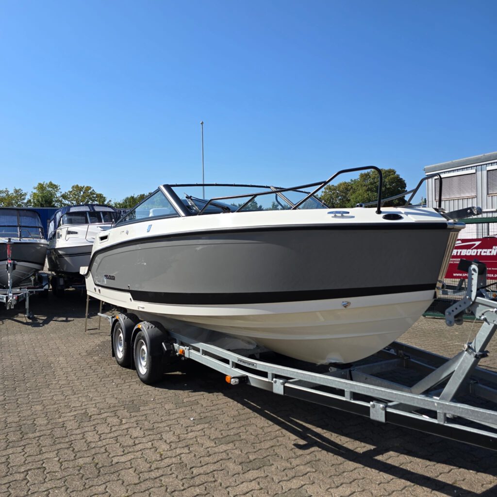 Ein weiß-graues Motorboot steht auf einem Bootsanhänger auf einem gepflasterten Platz vor einem klaren blauen Himmel.