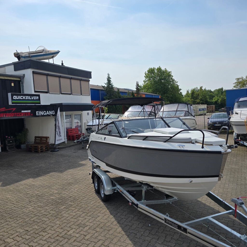 Ein weiß-graues Motorboot auf einem Anhänger vor einem Bootsverkaufsgebäude mit der Aufschrift Quicksilver und Eingang.