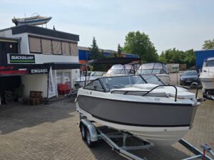 Ein weiß-graues Motorboot auf einem Anhänger vor einem Bootsverkaufsgebäude mit der Aufschrift Quicksilver und Eingang.