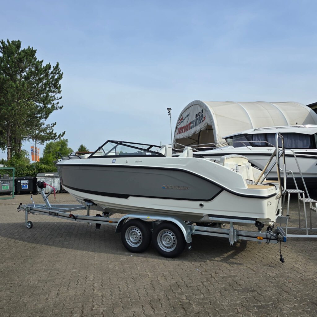 Ein graues Quicksilver-Motorboot steht auf einem Bootsanhänger vor einem großen Zelthangar auf einem gepflasterten Platz.